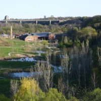 A green park with winding ponds and trees below a hilltop bridge and buildings.