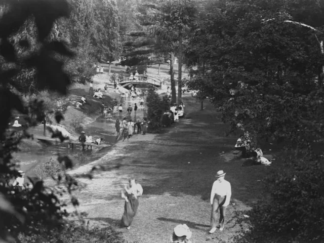 A black-and-white aerial view of people walking along a riverside path beneath trees.