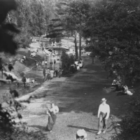 A black-and-white aerial view of people walking along a riverside path beneath trees.