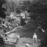 A black-and-white aerial view of people walking along a riverside path beneath trees.